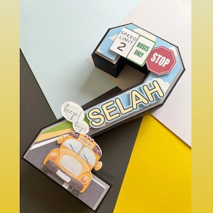May include: A three-dimensional papercraft number two decorated with a school bus theme. The number is black with yellow accents and features a yellow school bus, a stop sign, and a speed limit sign. The number is also decorated with the word "SELAH" in yellow letters.