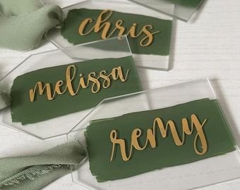 Op de afbeelding: Transparante acryl naamlabels met gouden letters en een groen geschilderde achtergrond. Elk label heeft een andere naam: Chris, Melissa, Remy en Kobe. Zachtgroen lint is aan de labels bevestigd, wat wijst op een bruiloft- of evenemententhema.