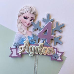 May include: A purple and blue cake topper featuring a character from the movie Frozen, a snowflake, the number 4, and the name Ayden. The topper is on a clear plastic stick.