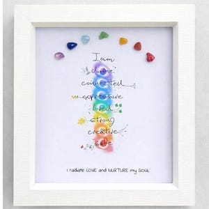 May include: A framed art print with a white frame. The artwork features a colorful illustration of the chakras, with gemstones and handwritten text. The text includes phrases like "I am divine, connected, expressive, loved, strong, creative, safe."