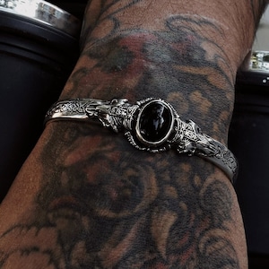 Op de afbeelding: Een zilveren armband met een sierlijk ontwerp en een zwarte edelsteen. De armband wordt gedragen op een getatoeëerde arm.