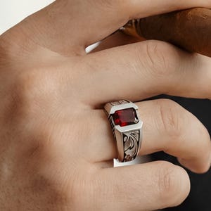 Può includere: Un anello d'argento con una gemma rossa rettangolare. L'anello presenta un design ornato e inciso sulla fascia. La persona tiene in mano un sigaro.