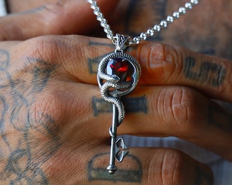 May include: A silver pendant necklace featuring a key design with a red gemstone heart and a snake wrapped around it. The necklace is worn on a silver chain.