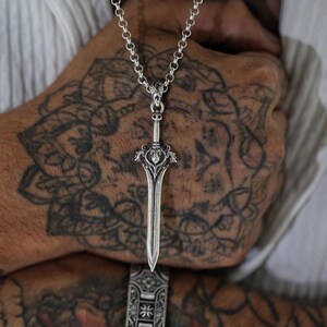 May include: A silver sword pendant necklace with a detailed design. The pendant is hanging from a silver chain.