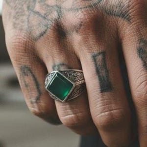 Op de afbeelding: Een zilveren ring met een vierkant geslepen, diepgroene steen in een zetting met ingewikkelde, getextureerde metaalpatronen aan de zijkanten. De ring wordt om een vinger gedragen en toont het gepolijste oppervlak van de steen en de band.
