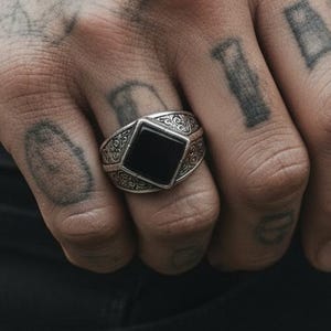 Può includere: Un anello d'argento con una pietra nera quadrata è indossato su un dito. L'anello ha un design ornato attorno alla pietra. La persona che indossa l'anello ha tatuaggi sulla mano e sul braccio. È visibile una cintura nera con fibbia argentata.