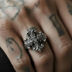 Op de afbeelding: Een zilveren ring met een sierlijk, symmetrisch bloemmotief. De ring heeft een gedetailleerd, krullend patroon met een centraal focuspunt. De ring wordt aan een vinger gedragen en toont zijn ingewikkelde vakmanschap en gotische stijl.