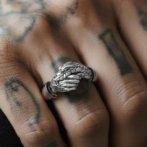 May include: A silver ring with a detailed snake design. The snake's head is the focal point, with intricate scales and a focused gaze. The ring is worn on a finger, highlighting the craftsmanship and the ring's aesthetic.