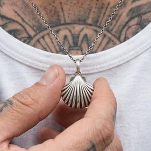 Può includere: Una collana in argento con un ciondolo a forma di conchiglia di capasanta con scanalature verticali. Il ciondolo pende da una sottile catenina d'argento testurizzata che poggia su una maglietta bianca e una giacca di pelle nera.