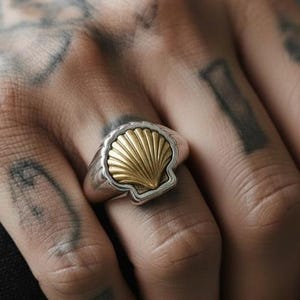 Puede incluir: Un anillo de plata con un diseño de concha dorada. El anillo presenta una forma de concha texturizada y detallada sobre una banda de plata pulida. Es una pieza llamativa, adecuada para el uso diario o ocasiones especiales.