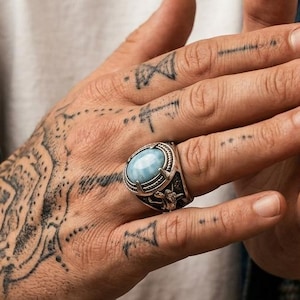 Könnte beinhalten: Ein silberner Ring mit einem großen, ovalen, hellblauen Edelstein. Der Ring hat aufwendige Seitendetails und wird an einer tätowierten Hand getragen. Die Person trägt eine Jeansjacke und ein weißes T-Shirt.