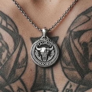 Op de afbeelding: Zilverkleurige ketting met een ronde hanger met een stierenkopontwerp. De hanger is gegraveerd met "MEMENTO TAURUS MORI". De ketting wordt gedragen op de borst van een persoon, met tatoeages zichtbaar op de achtergrond.
