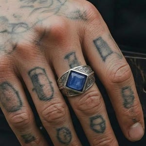 Puede incluir: Anillo de plata con una gema azul cuadrada. El anillo tiene un diseño ornamentado alrededor de la gema y en la banda. El anillo está en una mano con tatuajes.