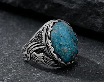 May include: A silver ring with an oval, turquoise-colored stone. The ring has an ornate design with detailed carvings around the stone and on the band, including wing-like patterns. The ring is set against a dark, textured background.