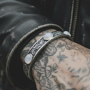Op de afbeelding: Een zilveren manchetarmband met een sierlijk, gegraveerd ontwerp en drie ovale, lichtgekleurde stenen. De armband wordt gedragen om een pols met gedetailleerde tatoeages, tegen een zwarte leren jas.