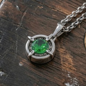 Puede incluir: Un collar con colgante de plata con una gema verde facetada, grande y redonda. El colgante tiene un diseño de brújula con las palabras "SOL JOT" alrededor de la gema. La cadena del collar también es plateada.