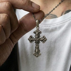 Może przedstawiać: Srebrzysty metalowy wisiorek w kształcie krzyża z misternymi, ozdobnymi zawijasami i detalami filigranowymi. Krzyż wisi na cienkim, pasującym srebrzystym metalowym łańcuszku, prezentując polerowane wykończenie z przyciemnionymi wgłębieniami dla głębi.
