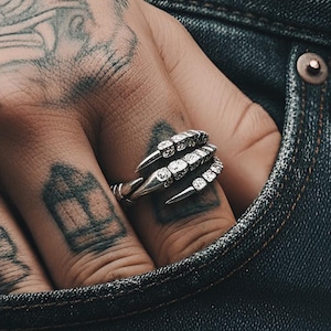 Puede incluir: Un anillo de plata con forma de garra y gemas transparentes. El anillo se lleva en un dedo, con la mano descansando en un bolsillo de mezclilla. El diseño del anillo presenta garras afiladas y puntiagudas que sostienen las gemas. La mano está tatuada.