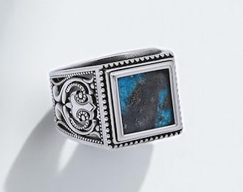 May include: A silver ring with a square, turquoise-colored stone. The ring has an ornate design on the sides and a textured border around the stone. The ring is set against a white background.