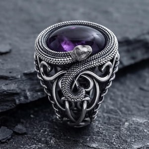 May include: A silver ring featuring a large, smooth, oval-shaped purple amethyst gemstone set in the center. The band is crafted with intricate metalwork depicting a coiled snake with textured scales wrapping around the stone, set against ornate scrollwork.