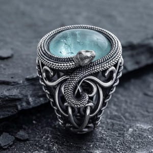 Puede incluir: Un anillo de plata con una piedra cabujón central de color azul claro lisa, rodeada por un diseño de serpiente enroscada con textura. La banda presenta intrincados patrones de volutas caladas que se extienden hacia abajo desde la serpiente.