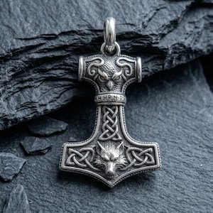 Può includere: Un ciondolo in metallo color argento a forma di martello, caratterizzato da una testa di gufo sulla parte superiore, una fascia centrale con il testo H H R N R, un intricato motivo a intreccio celtico e una testa di lupo alla base.