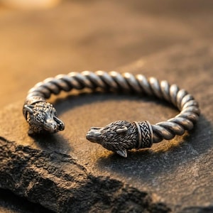 Può includere: Bracciale a polsino color argento con un design a corda intrecciata. Il bracciale presenta terminali dettagliati a forma di testa di lupo. Il bracciale è aperto, permettendo di indossarlo e toglierlo. Il bracciale è esposto su una superficie scura e strutturata.