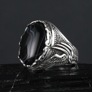 Peut inclure: Une bague en argent avec une pierre d'agate noire et des détails complexes en forme d'ailes. La bague est posée sur une surface noire.