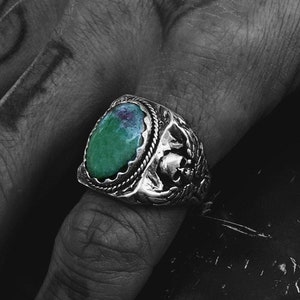 May include: A silver ring featuring a green gemstone set in an intricate design. The ring is worn on a hand with tattoos.