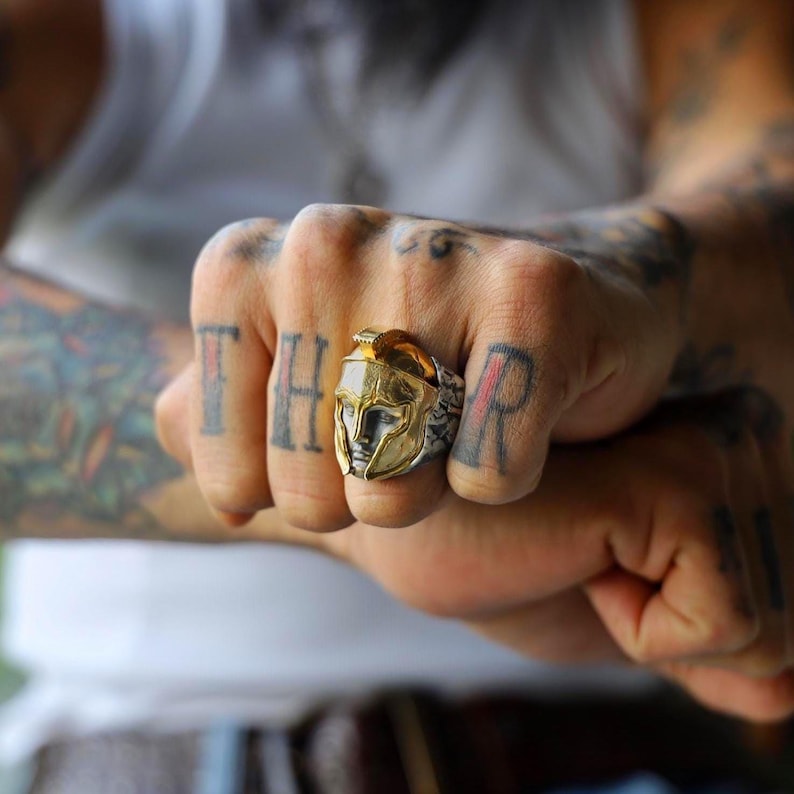 K&ouml;nnte beinhalten: Ein gold-silberner Ring mit einem Design eines spartanischen Helms. Der Ring wird an einer Hand mit T&auml;towierungen auf den Kn&ouml;cheln und dem Arm getragen. Der Helm hat detaillierte Merkmale und eine polierte Oberfl&auml;che.