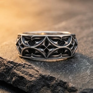 May include: A silver-toned metal ring featuring a repeating cutout pattern of pointed arches and diamond shapes. The recessed areas of the band are darkened, creating a high-contrast look that highlights the intricate, symmetrical metalwork design.