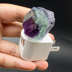 May include: A white night light with a purple and green fluorite crystal. The crystal is rough-hewn and sits atop the light fixture. The night light has a standard two-pin plug.