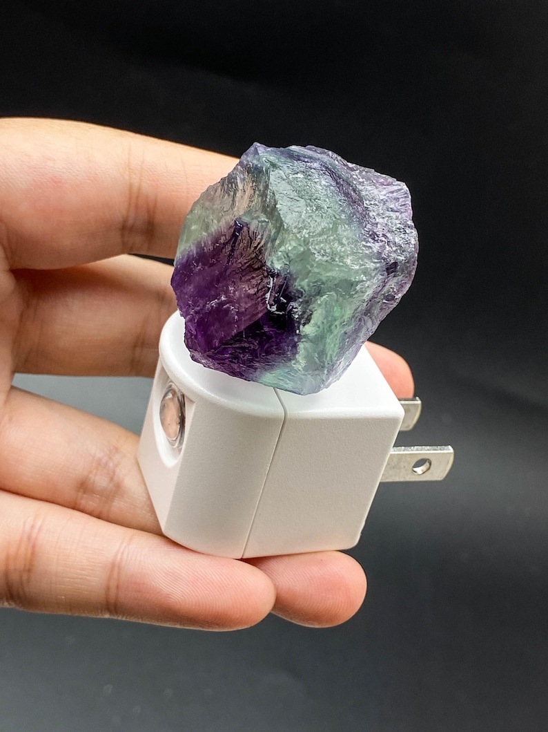 May include: A white night light with a purple and green fluorite crystal. The crystal is rough-hewn and sits atop the light fixture. The night light has a standard two-pin plug.