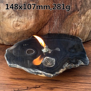 May include: A lit oil lamp set in a polished, black agate stone. The stone is irregularly shaped with a natural edge and a visible pattern. The flame is bright orange against the dark stone. The text in the image reads: 148x107mm, 281g.