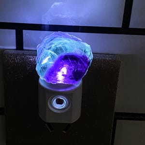 May include: A night light with a multi-coloured fluorite crystal. The crystal, with blue, purple, and clear tones, sits on a white plug-in base. The light provides a soft, ambient glow, suitable for bedrooms or hallways. The night light is a decorative item.