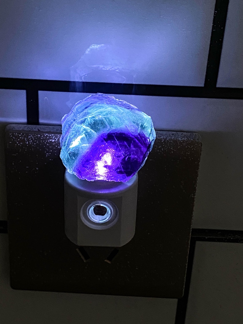 May include: A night light with a multi-coloured fluorite crystal. The crystal, with blue, purple, and clear tones, sits on a white plug-in base. The light provides a soft, ambient glow, suitable for bedrooms or hallways. The night light is a decorative item.