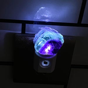 Peut inclure: Veilleuse avec un cristal de fluorite brut, affichant des nuances de bleu, violet et vert. Le cristal est éclairé, créant une douce lueur. La veilleuse est branchée sur une prise murale, offrant un éclairage d'ambiance.