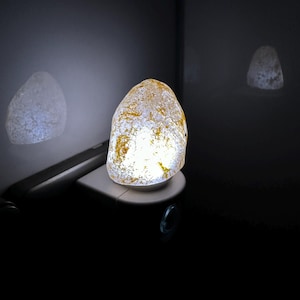 Peut inclure: Une veilleuse blanche avec une ampoule en verre clair et texturé qui émet une lueur chaude. L'ampoule a la forme d'une pierre arrondie et irrégulière.