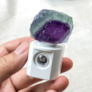May include: A white night light with a faceted design, topped with a rough-cut fluorite crystal. The crystal displays shades of purple and green. The night light has a circular light sensor.