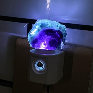 May include: A night light with a rough-cut fluorite crystal in blue and purple hues. The crystal sits on a white plug-in night light base, emitting a soft glow. The night light is plugged into a wall socket, providing ambient lighting.