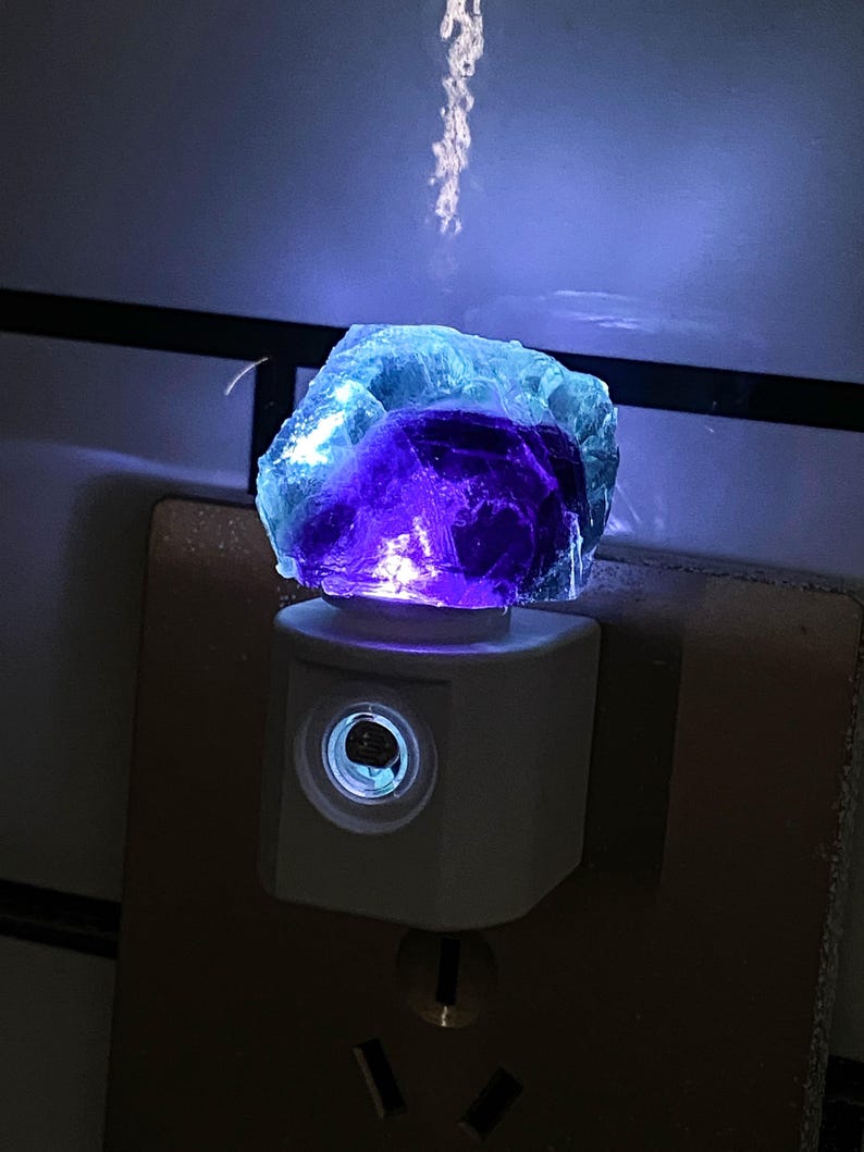 May include: A night light with a rough-cut fluorite crystal in blue and purple hues. The crystal sits on a white plug-in night light base, emitting a soft glow. The night light is plugged into a wall socket, providing ambient lighting.