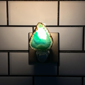 May include: A plug-in night light featuring a translucent, triangular slice of green agate stone. The stone has a dark, textured outer edge and glows with a bright white and emerald green light from within, mounted on a square base.