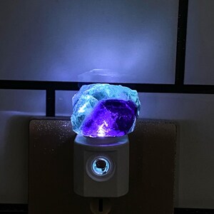 May include: A night light with a blue and purple fluorite crystal. The crystal sits on a white base that plugs into a wall socket. The light emits a soft glow, highlighting the crystal's natural colours.