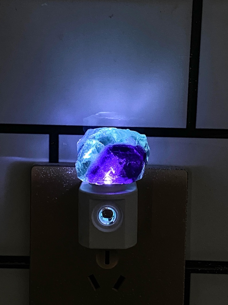 May include: A night light with a blue and purple fluorite crystal. The crystal sits on a white base that plugs into a wall socket. The light emits a soft glow, highlighting the crystal's natural colours.