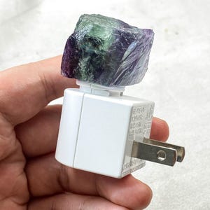May include: A white night light with a raw, multi-coloured fluorite crystal. The crystal features shades of green and purple. The night light has a standard two-pin plug.
