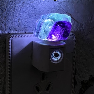 May include: A fluorite crystal night light, emitting a vibrant glow. The crystal, approximately 6 x 5 cm and weighing 70g, displays purple and blue hues. The night light is plugged into a wall socket.