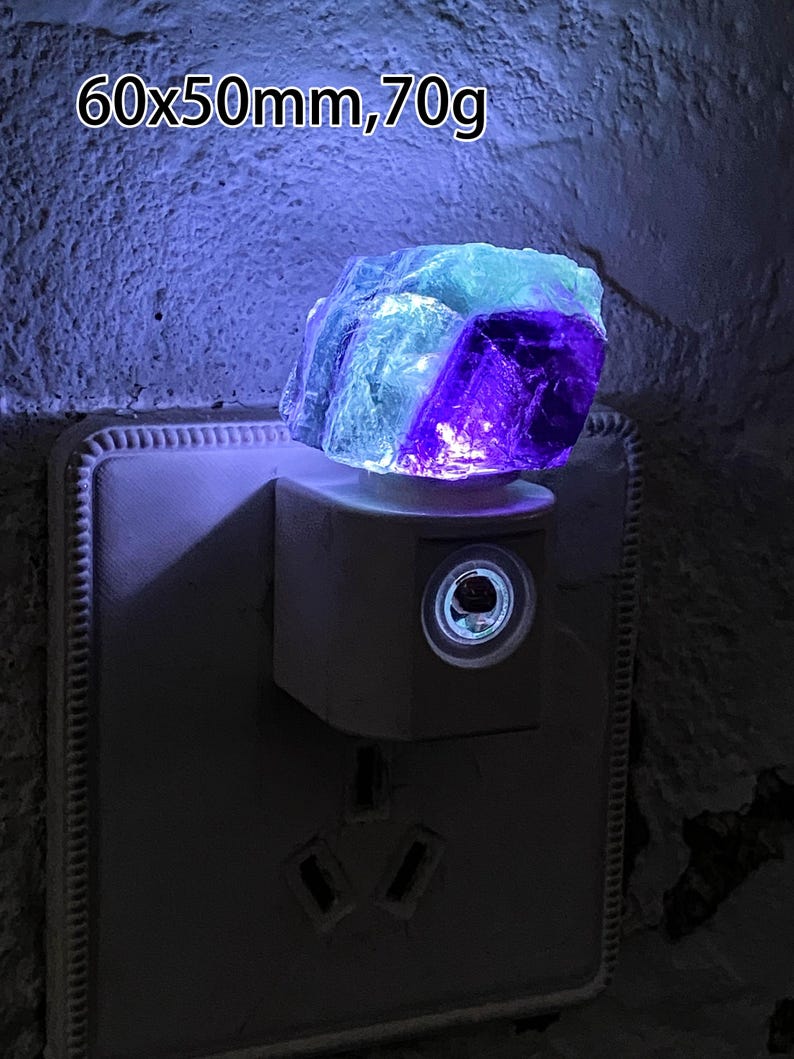 May include: A fluorite crystal night light, emitting a vibrant glow. The crystal, approximately 6 x 5 cm and weighing 70g, displays purple and blue hues. The night light is plugged into a wall socket.
