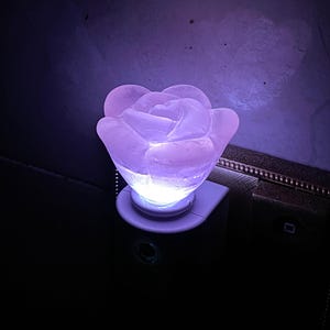 May include: A lavender-coloured, rose-shaped night light emitting a soft glow. The light is plugged into a wall socket. The rose is made of a translucent material, and the light source is visible within the petals, creating a gentle ambience.