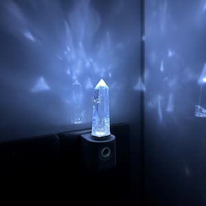 Op de afbeelding: Een helder kristallen nachtlampje dat in een stopcontact is gestoken. Het licht werpt een patroon op de muur erachter.