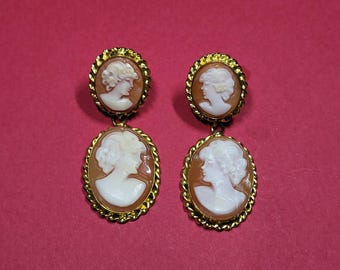 May include: A pair of drop earrings featuring oval-shaped cameos with white profiles of individuals set against a light brown background. Each cameo is encased in a gold-toned twisted rope metal frame, with a smaller circular cameo above a larger oval one.